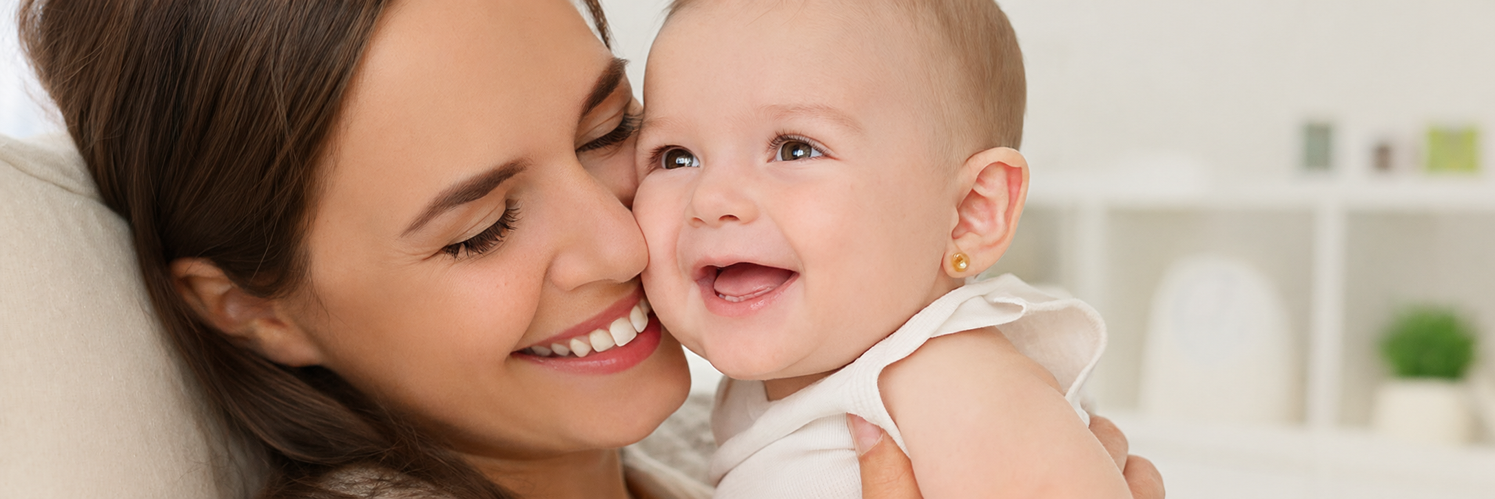 Mãe beijando o rosto do bebê, que usa um brinco pequeno e delicado na orelha